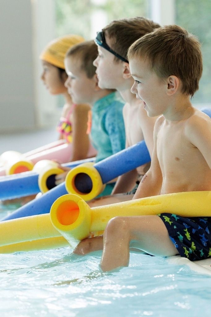 Kinder sitzen im Schwimmkurs nebeneinander am Beckenrand, jedes mit einer bunten Schwimmnudel, und hören aufmerksam zu