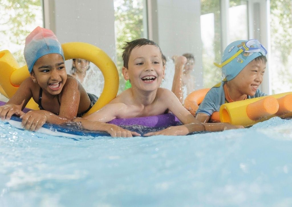 Lachende Kinder gleiten im Kinderschwimmkurs mit Schwimmbrett und Schwimmnudeln durchs Wasser