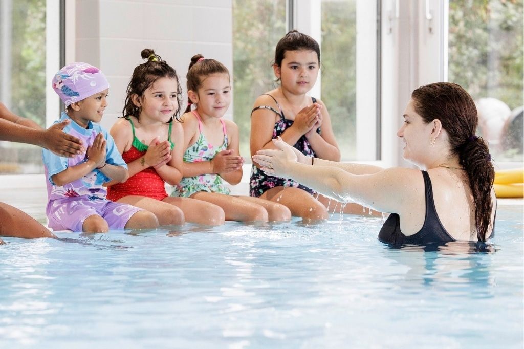 Schwimmtrainerin zeigt Kindern am Beckenrand im Wasser eine Übung, die Kinder machen die Armbewegung nach